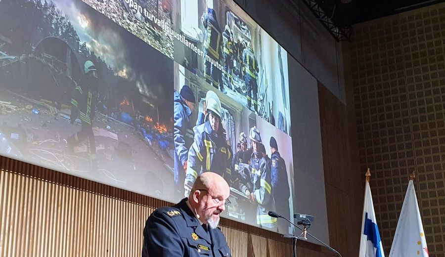 Pelastusylijohtaja Kimmo Kohvakka puhujanpöydän takana Suomen Sopimuspalokuntien Liiton Ajankohtaisseminaarissa Tampere-talolla. Taustalla kuvaesitys kansainvälisestä pelastustoimesta.